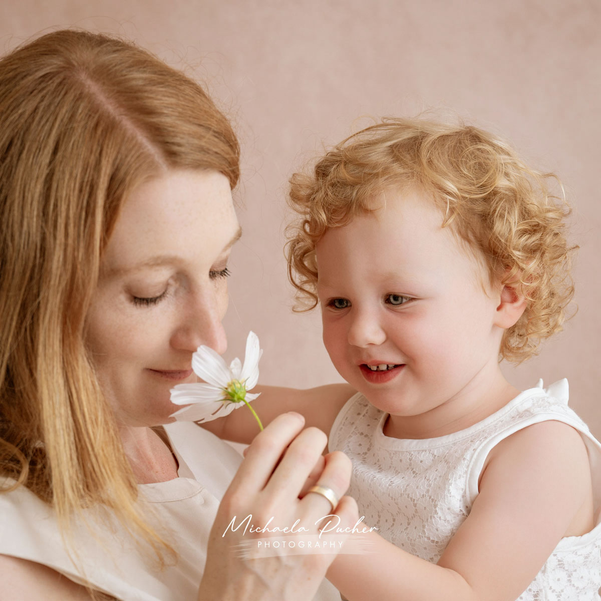 Mama hält eine weiße Blume und riecht daran, während ihre kleine Tochter sie lächelnd dabei beobachtet.